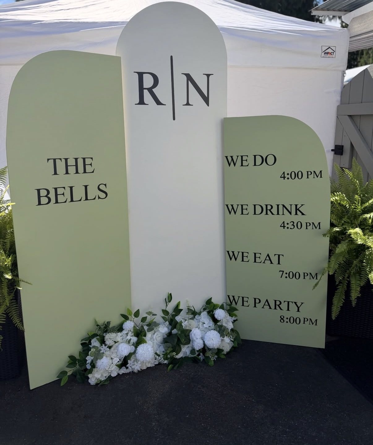 Sage and white arched signboards with monogram and an event timeline, dressed with white florals